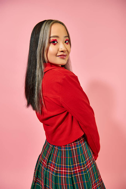 Sevgililer günü kırmızı ceketli Asyalı genç kadın pembe arka planda poz veriyor, kalp şeklinde göz makyajı yapıyor. - Fotoğraf, Görsel