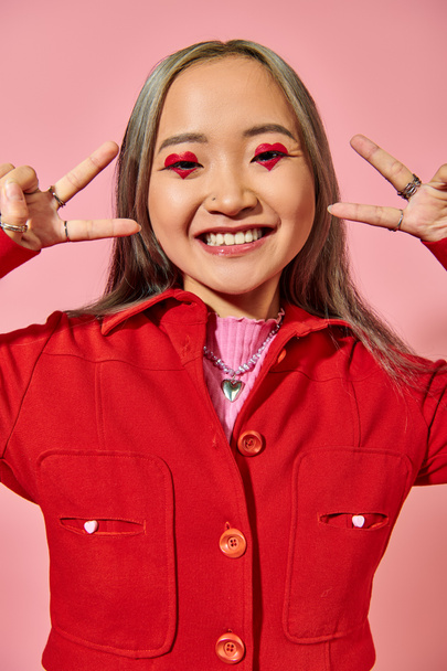boldog ázsiai fiatal nő piros kabát mutatja v jel rózsaszín háttér, szív alakú szem smink - Fotó, kép