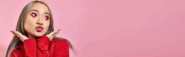 bonita asiática joven mujer con corazón forma ojo maquillaje puting labios y mirando hacia otro lado en rosa, bandera - Foto, Imagen