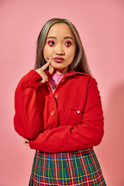zamyšlená asijská mladá žena s tvarem srdce oční make-up dotek tvář a odvrací pohled na růžové - Fotografie, Obrázek