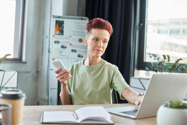 stylowa bigender osoba ze smartfonem pracującym na laptopie w pobliżu notebooka w nowoczesnym biurze - Zdjęcie, obraz