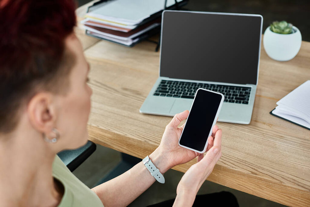 niebinarna osoba biznesowa trzymająca smartfona z pustym ekranem w pobliżu laptopa w miejscu pracy w biurze - Zdjęcie, obraz
