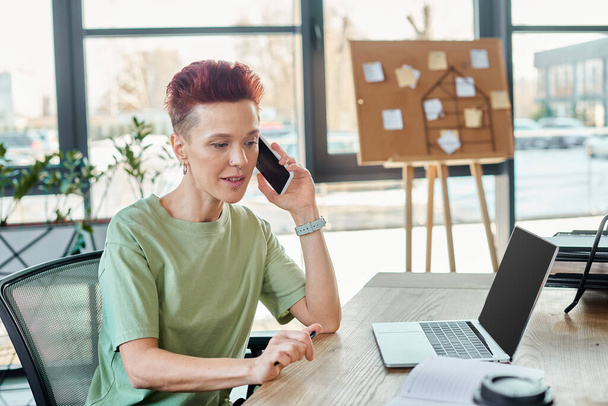 non-binary business person talking on mobile phone near laptop with blank screen in modern office - Photo, Image