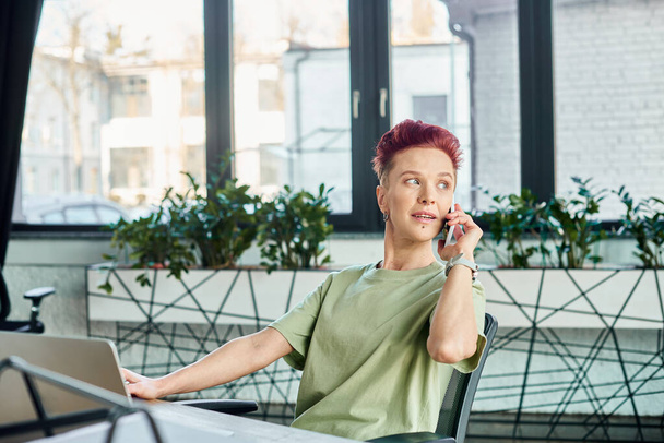 bigender business person talking on mobile phone near laptop and looking away in modern office - Photo, Image