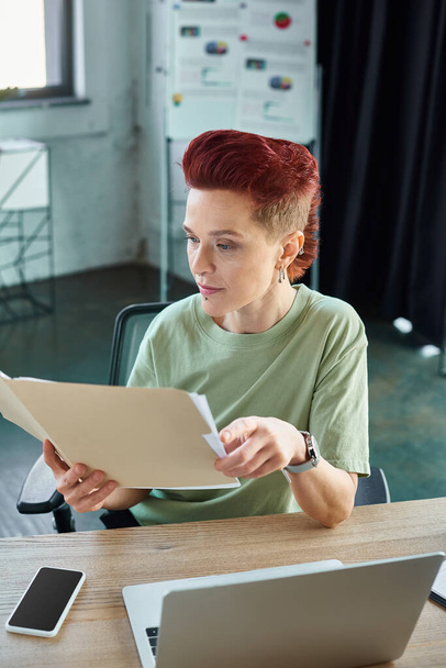 gerente queer elegante trabalhando com documentos perto de laptop e smartphone com tela em branco no escritório - Foto, Imagem