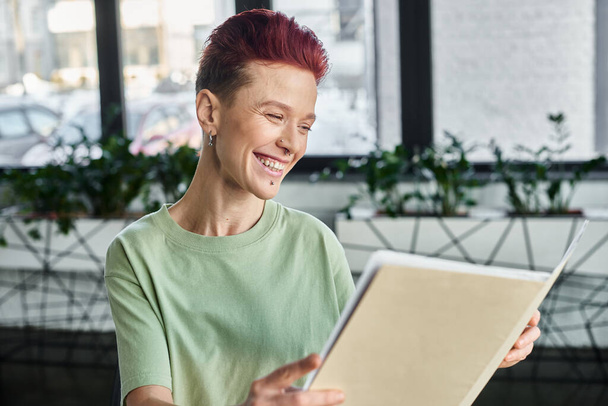 joyful queer person with short hair looking  at folder with documents in contemporary office - Photo, Image