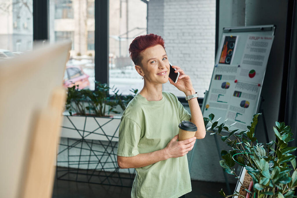 heureux queer personne avec du café pour aller dans la tasse en papier parler sur téléphone mobile dans le bureau moderne - Photo, image