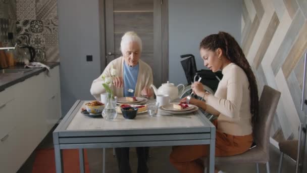 Zoom in shot di donna anziana e assistente sociale femminile che discutono mentre bevono tè e mangiano dolci insieme a casa - Filmati, video