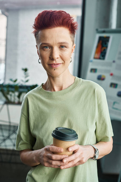 smiling non-binary person with takeaway drink in paper cup looking at camera in modern office - Photo, Image