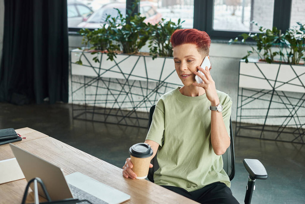 positive queer person with coffee to go in paper cup talking on mobile phone near laptop in office - Photo, Image