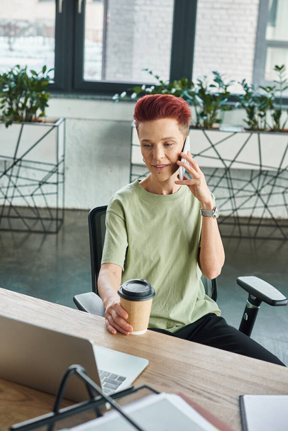 persona queer positivo con café para ir en taza de papel hablando en el teléfono móvil cerca de la computadora portátil en la oficina - Foto, Imagen