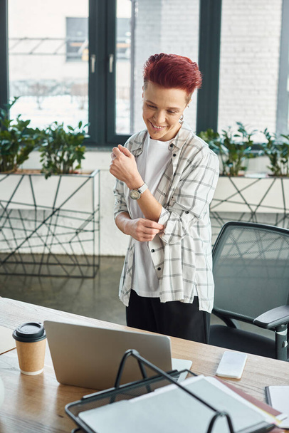 fröhliche Bigender-Managerin in lässiger Kleidung, die am Arbeitsplatz steht und im Büro auf Laptop schaut - Foto, Bild