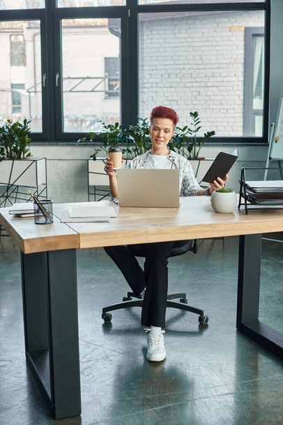 stylish non-binary person with coffee to go and notebook sitting near laptop in modern office - Photo, Image