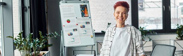 szczęśliwy bigender osoba stojąca w miejscu pracy w nowoczesnym biurze i patrząc w aparat fotograficzny, baner - Zdjęcie, obraz