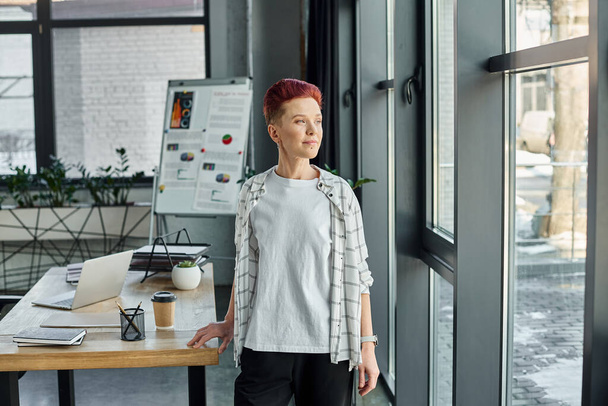thoughtful queer person in casual attire standing near windows and looking away in modern office - Photo, Image