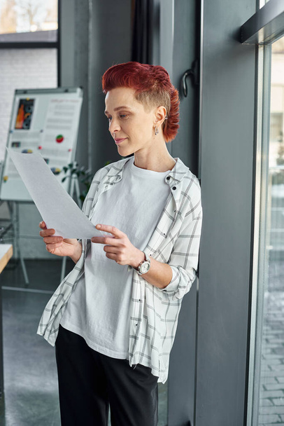 persona no binaria reflexivo en traje casual mirando los documentos, mientras que de pie en la oficina moderna - Foto, Imagen