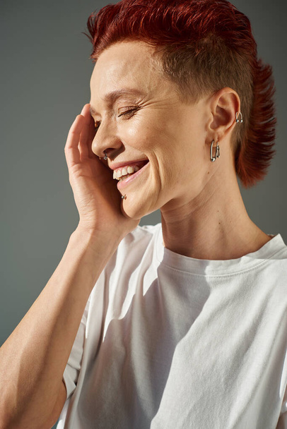 redhead bigender person with piercing smiling with closed eyes and hand near face on grey, portrait - Photo, Image