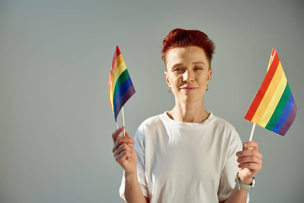 pelirroja persona no binaria en camiseta blanca de pie con pequeñas banderas de colores del arco iris en gris - Foto, Imagen