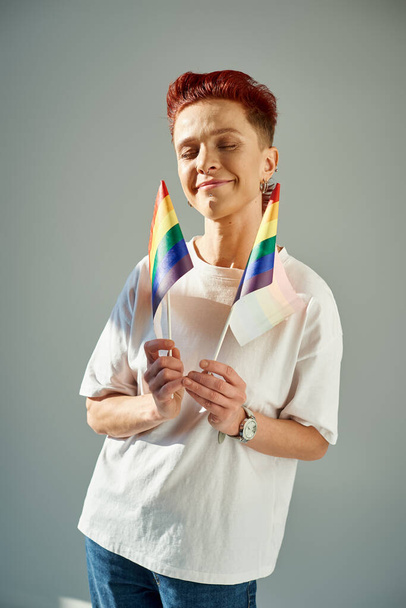 rossa persona queer in t-shirt bianca con piccole bandiere LGTB e sorridente con gli occhi chiusi sul grigio - Foto, immagini