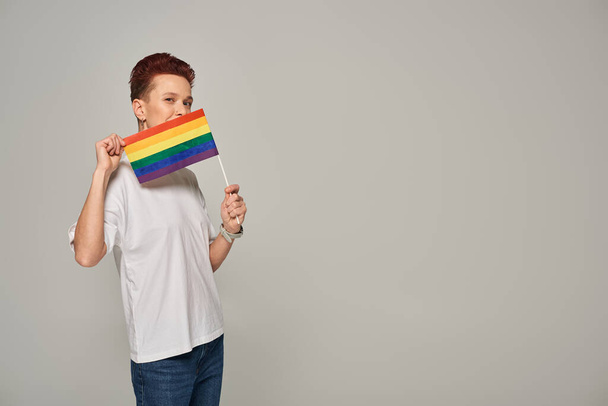 pelirroja modelo queer en camiseta blanca posando con pequeño LGBT plano cerca de la cara mirando a la cámara en gris - Foto, imagen
