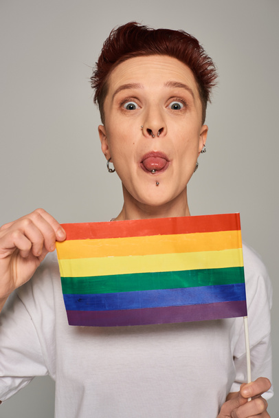 ruda queer model z kolczykiem i mały LGBT płaskie wystające i patrząc w aparat fotograficzny na szary - Zdjęcie, obraz