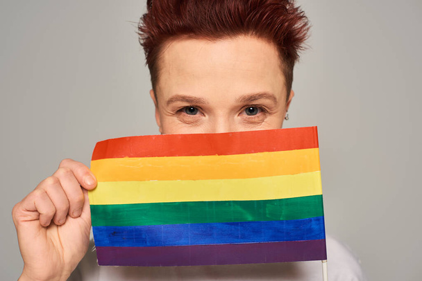 alegre ruiva queer pessoa obscurecendo rosto com pequena bandeira LGBT e olhando para a câmera no cinza - Foto, Imagem