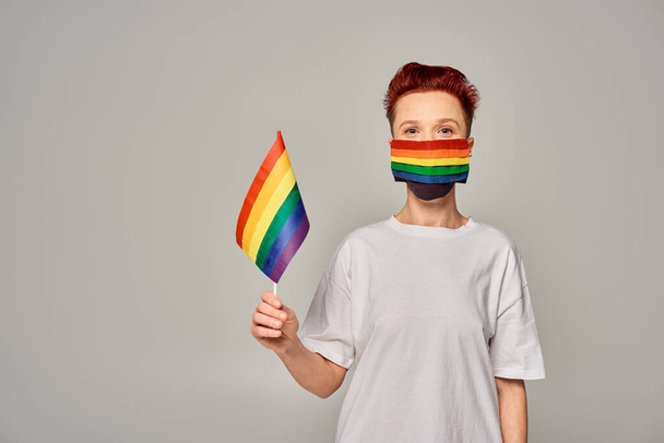 pelirroja queer persona en blanco camiseta y arco iris colores máscara médica con pequeña bandera LGBT en gris - Foto, imagen