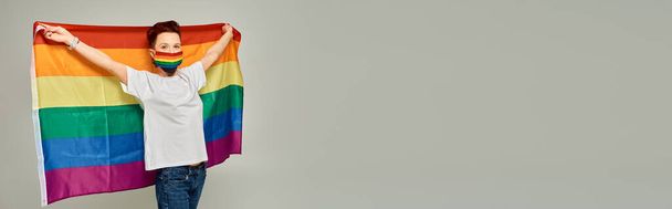 κοκκινομάλλα queer μοντέλο σε χρώματα ουράνιο τόξο ιατρική μάσκα κρατώντας σημαία ΛΟΑΤ σε γκρι, οριζόντια banner - Φωτογραφία, εικόνα