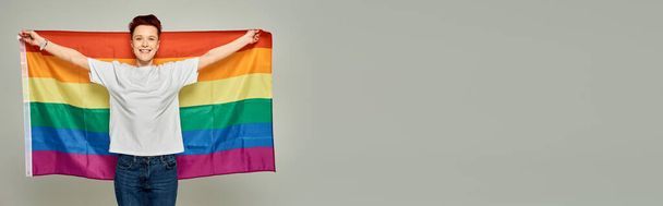 Gri arka planda LGBT bayrağı taşıyan beyaz tişörtlü neşeli kızıl saçlı kişi, afiş. - Fotoğraf, Görsel
