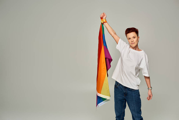 vážná zrzka bigender osoba v bílém tričku a džíny stojící s vlajkou LGBT na šedém pozadí - Fotografie, Obrázek
