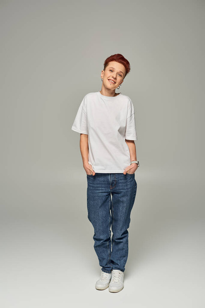full length of happy redhead queer person in white t-shirt and jeans with hands in pockets on grey - Photo, Image
