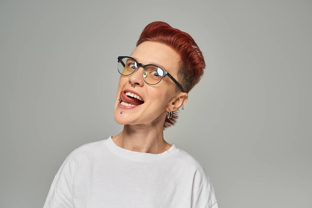 funny and cheerful non-binary person in white t-shirt and eyeglasses sticking out tongue on grey - Photo, Image