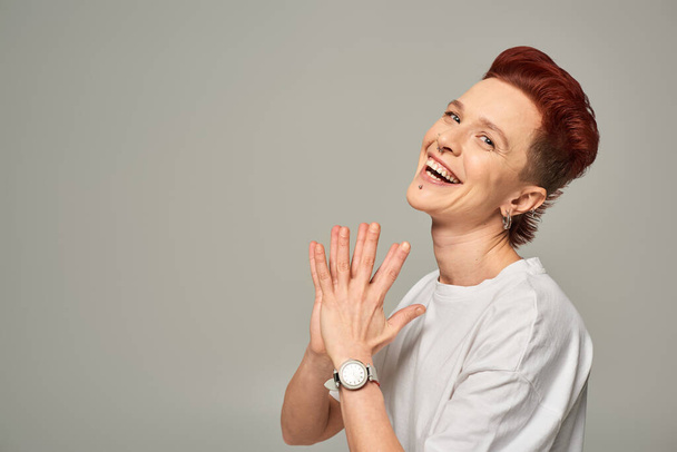 cheerful redhead bigender person in white t-shirt laughing and looking at camera on grey, happiness - Photo, Image