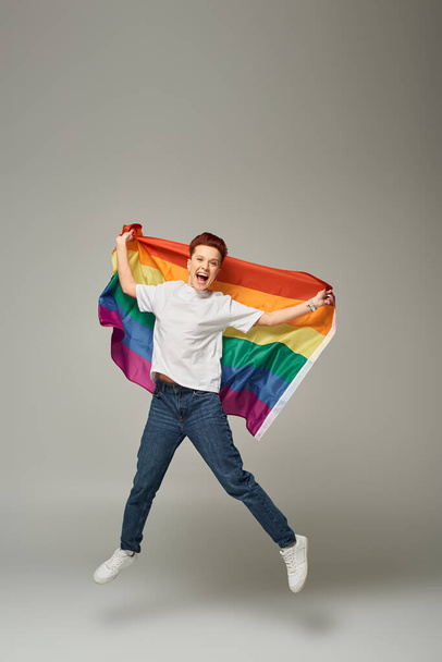 persona queer muy alegre en camiseta blanca y pantalones vaqueros saltando y levitando con bandera LGBT en gris - Foto, imagen