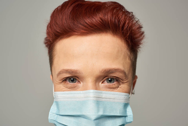 zbliżenie portret rudej osoby queer ze szczęśliwym spojrzeniem w masce medycznej patrząc na kamerę na szarości - Zdjęcie, obraz
