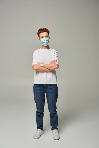 pelirroja positiva queer persona ganar blanco camiseta y máscara médica de pie con los brazos cruzados en gris - Foto, Imagen