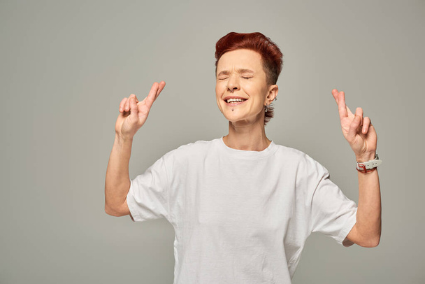 napięty bigender osoba w białym t-shirt skrzyżowanie palce na szczęście stojąc z zamkniętymi oczami na szary - Zdjęcie, obraz