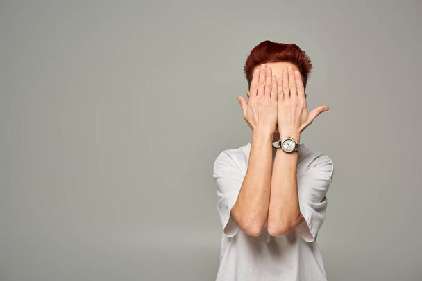 redhead bigender person in white t-shirt obscuring face with hands while standing on grey backdrop - Photo, Image