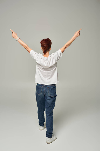 back view of redhead queer person in white t-shirt and jeans pointing aside with fingers on grey - Photo, Image