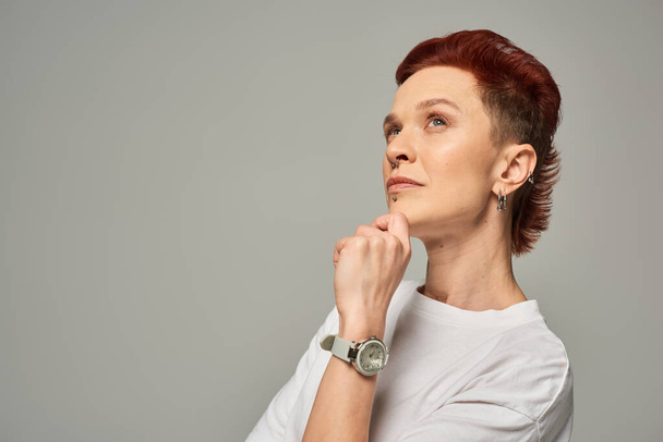 thoughtful redhead non-binary person holding hand near chin and looking away on grey backdrop - Photo, Image