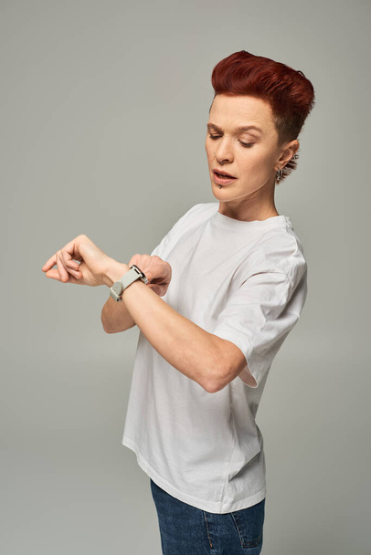 persona pelirroja grande grave en camiseta blanca comprobar el tiempo en reloj de pulsera sobre fondo gris - Foto, Imagen