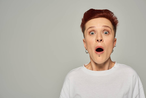 σοκαρισμένη κοκκινομάλλα queer άτομο σε λευκό t-shirt στέκεται με ανοιχτό το στόμα και κοιτάζοντας κάμερα σε γκρι - Φωτογραφία, εικόνα