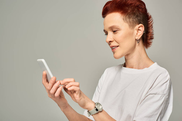 χαμογελαστή κοκκινομάλλα queer πρόσωπο σε λευκό T-shirt μηνυμάτων στο κινητό τηλέφωνο, ενώ στέκεται σε γκρι - Φωτογραφία, εικόνα