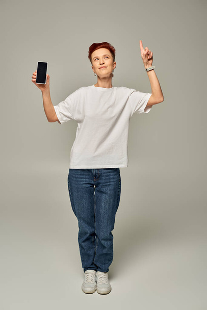 queer person holding smartphone with blank screen while looking up and pointing with finger on grey - Photo, Image