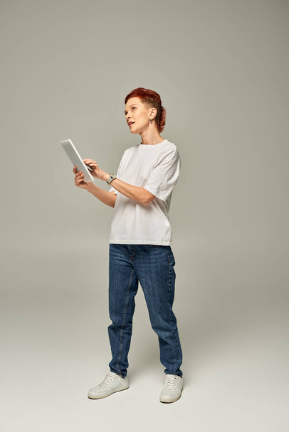 przemyślana osoba niebinarna w białej koszulce z wykorzystaniem tabletu cyfrowego i odwracająca wzrok na szarym tle - Zdjęcie, obraz