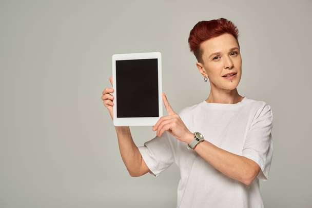 rudowłosa osoba niebinarna w białej koszulce pokazująca cyfrową tabletkę z czystym ekranem na szarym tle - Zdjęcie, obraz