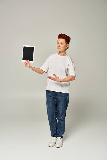 рыжеволосый небинарный человек в белой футболке показывает цифровой планшет с пустым экраном на сером фоне - Фото, изображение