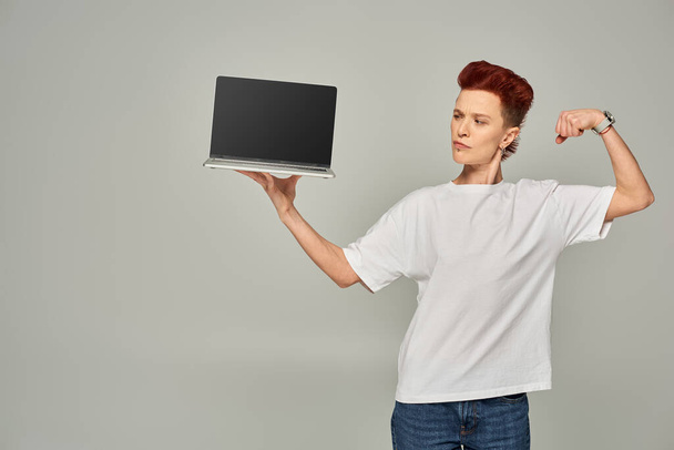 Freelancer queer confiado mostrando músculos mientras sostiene el ordenador portátil con pantalla en blanco sobre fondo gris - Foto, Imagen