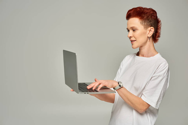 freelance queer rousse en t-shirt blanc réseautage sur ordinateur portable tout en se tenant sur fond gris - Photo, image