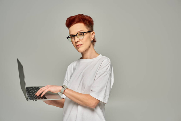 rothaarige queere Freiberuflerin in weißem T-Shirt und Brille vernetzt auf Laptop vor grauem Hintergrund - Foto, Bild